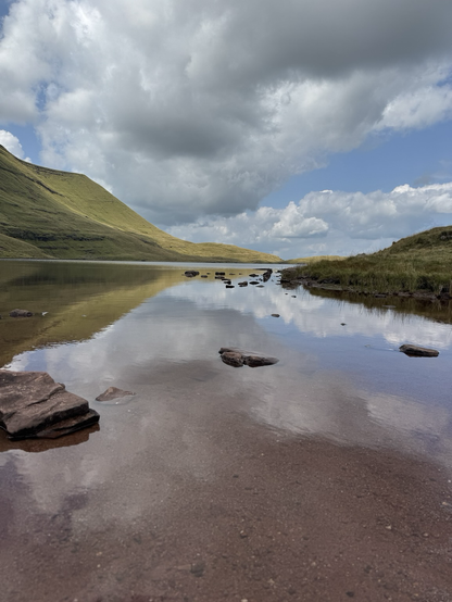 Green hill slopes down from the left to a glassy lake which is mirroring the blue sky with clouds above.