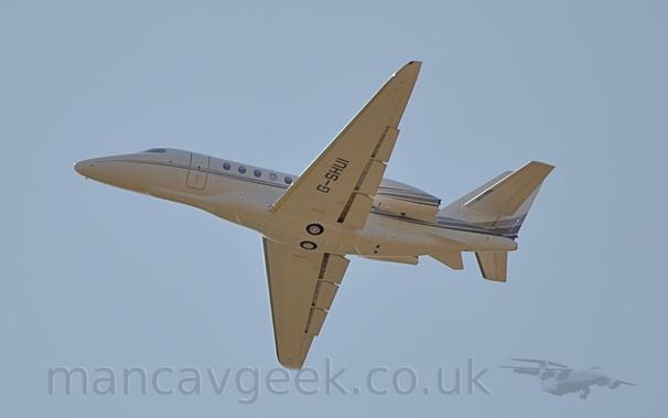 Low side view of a twin engined BizJet flying from right to left at a very low altitude, with the undercarriage tucked away, flaps extended from behind the wings, and the nose raised sharply, suggesting that it has just taken off.
The plane is mostly white, with a 3-tone running along the fuselage from the nose, as well as along the bottom of the engine pods mounted on the side of the rear fuselage.
The black registration "G-SHUI" is on the underside of the left wing.
Pale blue sky fills the rest of the frame.