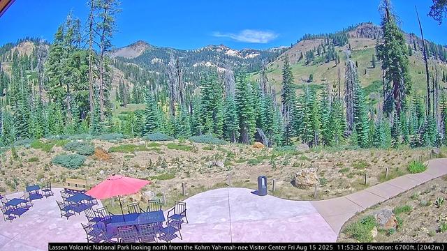 The field of view from this camera in Lassen Volcanic National Park has been changed. The view has been expanded. A seating area with tables, chairs, and a red sun umbrella appears in the foreground. Grass, rocks, and a very nice variety of trees appear in the center of the image. Distant peaks, some forested, can be seen in the background, under a beautiful blue sky, which is hosting a single white cloud in this particular image.