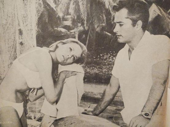 Ursula Andress en un entorno tropical, sentada y luciendo un bikini blanco, mientras observa a un hombre a su lado que viste una camisa blanca. La escena refleja un ambiente de glamour y sensualidad, evocando la era dorada del cine.