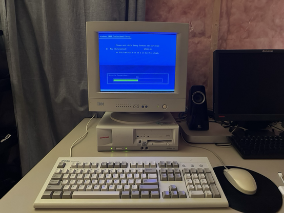 A vintage PC setup shows a Compaq Deskpro EN desktop computer placed beneath an IBM CRT monitor, which is currently displaying the Windows 2000 Professional Setup screen. The setup is in the process of formatting a partition labeled C: New (Unformatted) with a size of 57,239MB, and the progress bar indicates it’s 51% complete. The familiar blue setup screen states: “Please wait while Setup formats the partition…”  In front of the computer is a classic Dell QuietKey keyboard with gray and white keys, along with a generic beige two-button mouse resting on a black mousepad. A pair of Logitech desktop speakers is visible behind the keyboard on the right, next to a powered-off Lenovo ThinkVision monitor and another keyboard.  The scene is set against a backdrop of unfinished walls with visible pink insulation, and the workspace has a cozy, retro-tech atmosphere. The CRT’s glow adds a nostalgic touch, capturing a moment familiar to anyone who installed Windows operating systems in the late 1990s or early 2000s.