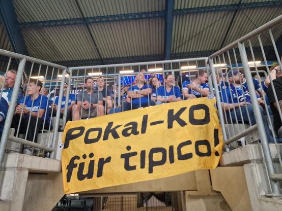 Oranger Banner mit der Aufschrift: "Pokal-KO für Tipico!". Davor sind Fans des Vereins Arminia Bielefeld zu sehen.