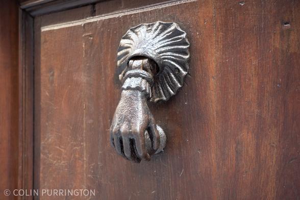 Gracile metal hand clutching a ball that can be moved to strike a plate on a wood door.