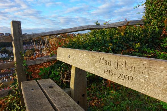 A weathered wooden bench sits on a grassy hill overlooking a town and the sea. The backrest of the bench has an inscription carved into it, which reads "Mad John ? 1945 ~ 2009". A rustic wooden fence runs behind the bench, and the area is surrounded by lush green and autumnal brown foliage. The sun is low in the sky, casting a warm light over the scene, and a few clouds are visible against the blue sky. The photo is taken from a low angle, focusing on the bench and the inscription.