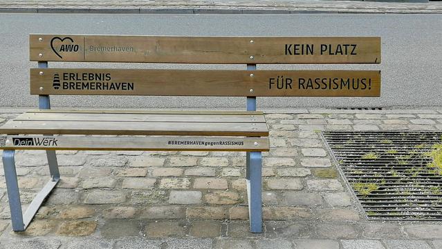 Bank mit Sitzfläche ind Lehne als Holzlatten. Am rechten Rand der Lehne steht „ Kein Platz für Rassisten“. Unter dem Schriftzug fehlt die Sitzfläche.