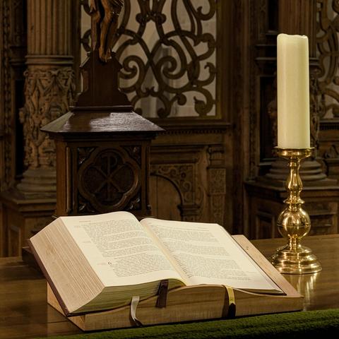 Open book on a wooden lectern with intricate carvings, placed on a wooden surface. A tall, unlit white candle stands in a gold candlestick nearby. Background features ornate wood detailing and scrollwork.
###
Offenes Buch über ein hölzernes Rednerpult mit aufwendigen Schnitzereien, die auf einer Holzoberfläche platziert sind. Eine große, unbeleuchtete weiße Kerze steht in einem goldenen Kerzenständer in der Nähe. Hintergrund zeigt kunstvolle Holzdetails und Scrollwork.
