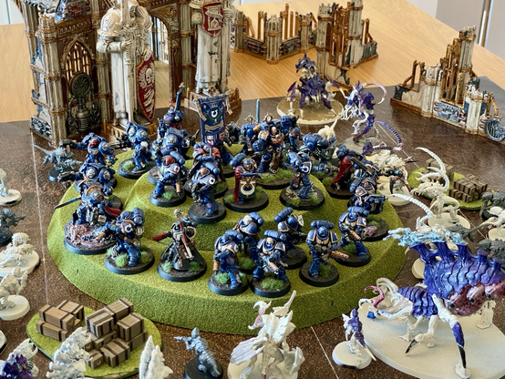 Many ultramarines models standing on a hill, facing outwards, and surrounded by tyranid models, in a «last stand» scene. In the center at the top of the hill is a model carrying the ultramarines standard. Ultramarines models are painted. Most tyranid models are unpainted.