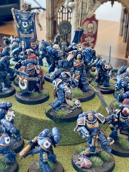 Many ultramarines models standing on a hill, facing outwards, and surrounded by tyranid models, in a «last stand» scene. In the center at the top of the hill is a model carrying the ultramarines standard. Ultramarines models are painted. Most tyranid models are unpainted. Closeup of the top of the hill.