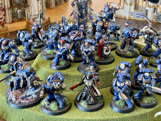 Many ultramarines models standing on a hill, facing outwards, and surrounded by tyranid models, in a «last stand» scene. In the center at the top of the hill is a model carrying the ultramarines standard. Ultramarines models are painted. Most tyranid models are unpainted. Different angle.