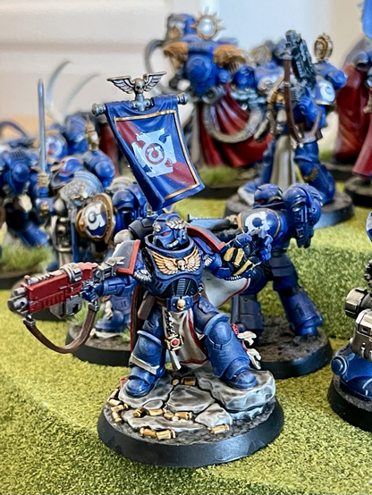 Many ultramarines models standing on a hill, facing outwards, and surrounded by tyranid models, in a «last stand» scene. In the center at the top of the hill is a model carrying the ultramarines standard. Ultramarines models are painted. Most tyranid models are unpainted. Captain Centos closeup.