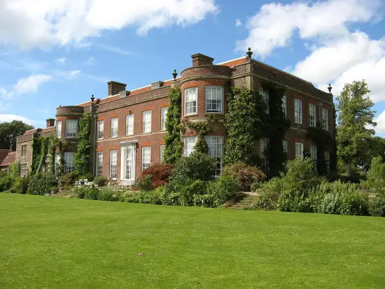 The image depicts a large, elegant brick house surrounded by lush greenery. It features multiple windows, some of which are adorned with climbing plants. The building has a prominent garden area in front, with a well-kept lawn and various shrubs and trees. The sky is partly cloudy, indicating a pleasant day.

Image Credits: Wikimedia / Rwendland / CC BY-SA 3.0