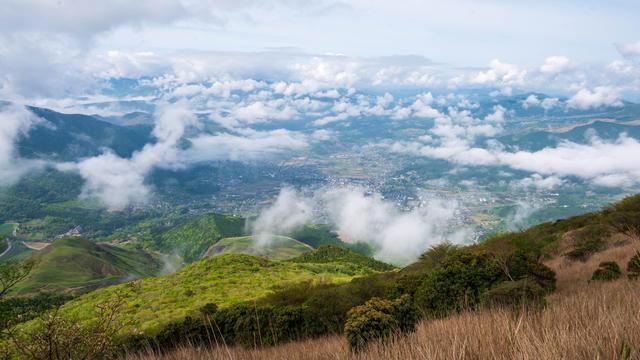 A panoramic view of a lush green valley surrounded by mountains, partially covered by clouds. The scene captures a mix of greenery, fields, and a small village nestled in the landscape beneath a blue sky with scattered clouds.