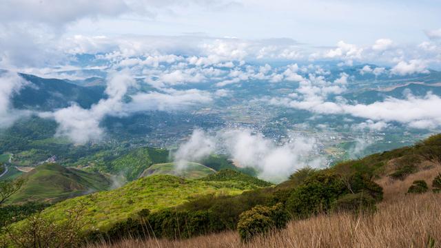 A panoramic view of a lush green valley surrounded by mountains, partially covered by clouds. The scene captures a mix of greenery, fields, and a small village nestled in the landscape beneath a blue sky with scattered clouds.