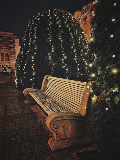 A vertical, eye-level shot captures a carved stone bench on a brick walkway at night, nestled between two large evergreen shrubs decorated with strings of warm, white lights. In the background, buildings are dimly lit and several people walk along the path.