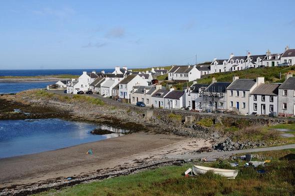 Portnahaven. The image shows a view down across the head of a tidal inlet with a boat pulled up on the grass. A road leads along the far side of the inlet with white houses and cottages on its far side. More houses are placed along a ridge above and behind the first row. The sky is blue.