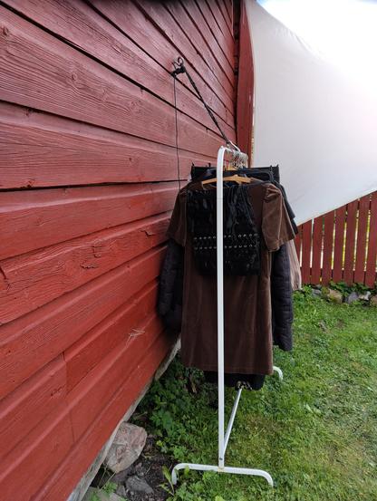 Vaaterekissä roikkuu vaatteita henkareista. <Rekki on punaista lautaseinää vasten. Seinässä on koukku, josta lähtee naru, jonka päässä on semmoinen kiinnityshakanen. Nyt se on kiinnitetty rekin ylätankoon.