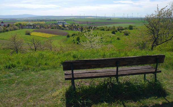 Eine Landschaftsaufnahme. Das Foto wurde hinter einer Bank, die auf einem Berg oberhalb von einem Dorf steht, aufgenommen. Im Hintergrund sieht man den Harz und Felder und Windkraftanlagen.