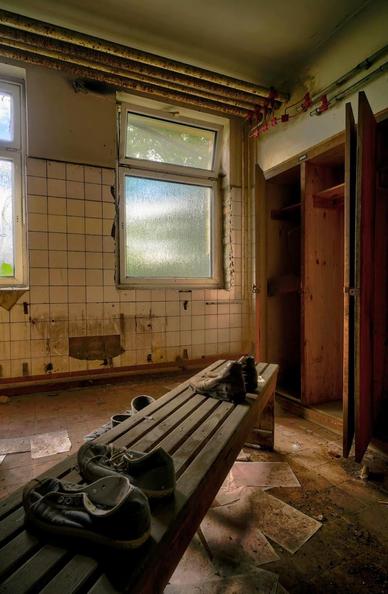 Foto in einem Souterrain-Raum mit Milchglasscheiben. eine hölzerne Umkleidebank steht diagonal im Raum, darauf 2 paar alte Schuhe. 
Verschmutzte Zeitschriften-Seiten am Boden. Rechts offene Holz-Spinde. Der Heizkörper vor der weiß gekachelten Wand fehlt. Unter der Decke offen verlegte Rohre.
