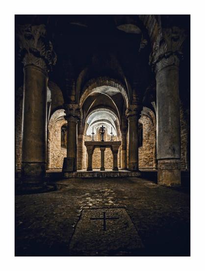 This is a photograph of is an ancient stone chapel with a vaulted ceiling, (La Cripta di San Zama) it has arched columns and a central altar with a cross on top of it. The dim lighting highlights the rough textures of the brick walls and stone floor, creating a somber, historic atmosphere. In the foreground is a cross on the floor and almost points you towards the altar.