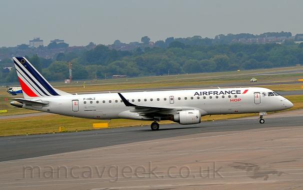 Side view of a twin engined jet airliner taxiing from left to right.
The plane is mostly white, with blue "Air France" titles on the upper forward fuselage, and smaller "Hop" titles underneath.
The black registration "F-HBLZ" is on the upper rear fuselage, while the last 2 letters "LZ" are on the open nose-wheel doors.
3 diagonal blue stripes are on the tail, getting narrower the further back they are, with a fourth, red stripe at the back, the same width as that at the front.
Grey concrete apron and black concrete taxiway fill the foreground, with grass, taxiways, and a runway in the background, leading up to trees in the distance, under grey skies.