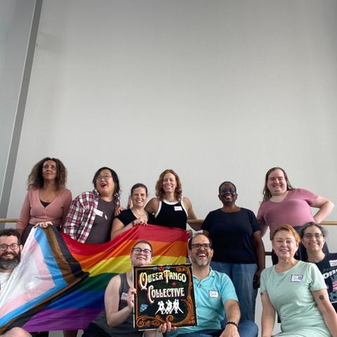 11 people smile, gathered around a Pride flag and a sign that says “queer tango collective”