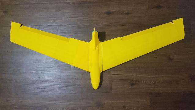 A photo of a yellow flying wing RC plane body on a wooden floor.