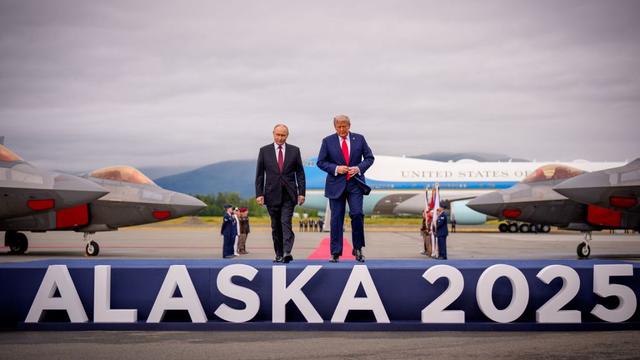 Two men in suits walk on a platform with large white letters spelling "ALASKA 2025" at an airbase with fighter jets and an Air Force One plane in the background under a cloudy sky.