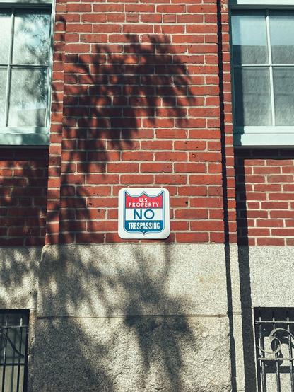 Red brick wall with two windows and a “U.S. PROPERTY NO TRESPASSING” sign. A band of gray stone runs along the base, and soft shadows of tree branches stretch across the facade.