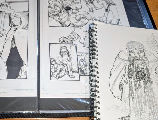 A sketchbook lays on top of an open art portfolio. The sketchbook shows a half-inked drawing of an angry looking dwarf king with a bajillion braids holding an art staff. Two black and white comic pages are visible in the open portfolio. They include images of a masked woman, a dwarf in armor, and three young boys.
