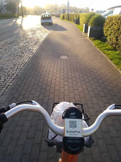 Blick über den Lenker des Leihrads "Schleifee" auf den gepflasterten Radweg, im Fahrradkorb Brötchen, links vorne parkt ein Mietauto, daneben die Straße, die Sonne beleuchtet von links oben das Bild.