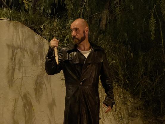 Un hombre calvo y barbudo, con una barba oscura y bien cuidada, aparece de pie en un entorno oscuro, probablemente de noche. Viste una gabardina larga de cuero oscuro sobre una camiseta blanca. En su mano derecha, sostiene firmemente una estaca, levantado a la altura de su pecho. Su mirada es seria y se dirige hacia el lado derecho de la imagen. Detrás de él, a la izquierda, hay un muro de color claro y texturizado, mientras que a la derecha se distingue una densa vegetación oscura. La escena está iluminada por una luz artificial que crea contrastes y sombras.