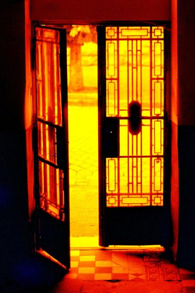 The image depicts an open door with intricate glass panels, allowing bright yellow light to flood into a dimly lit interior space. The door is framed by dark wood, with a geometric pattern of rectangles and lines on the glass. The left door is slightly ajar, while the right door is fully open, revealing a pathway outside. The floor inside the doorway features a checkered pattern of light and dark tiles, transitioning to a brighter, sunlit area outside. The warm, yellow light creates a striking contrast with the darker interior, highlighting the door's design and the transition from inside to outside.