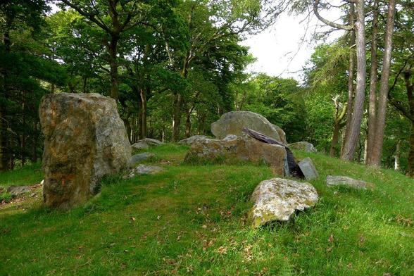 A group of megaliths set on top of a grassy mound in woodland. A standing stone on the left, with slabs forming a  chamber in the centre. It's a fine morning, and sunlight is filtering through the trees.
