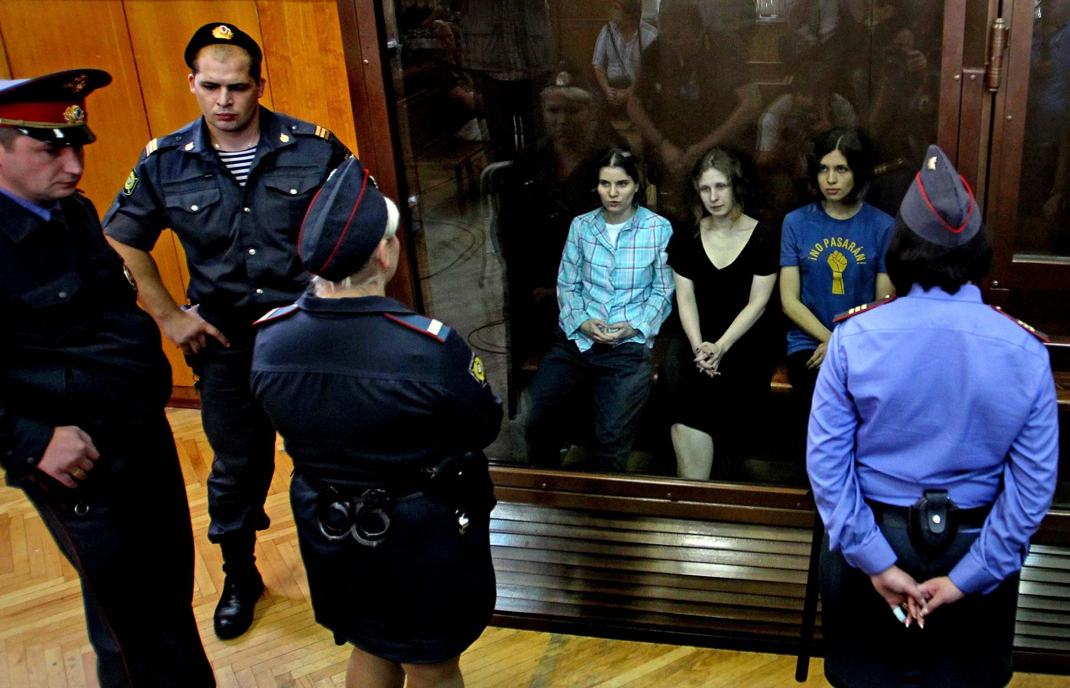 Maria Alyokhina, Nadezhda Tolokonnikova, and Yekaterina Samutsevich in court hearing their sentence. They are three white women sitting behind plexiglass and being guarded by several military personnel.