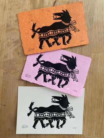 Three postcard-sized linocut prints : a stylized wolf with a sun in its jaws, wearing a cape adorned with stars and geometric shapes. Under the cape are six human legs, revealing that the wolf is a costume worn by three persons in line. The picture is printed in black ink on handmade orange and pink papers as well as off-white Fabriano paper.