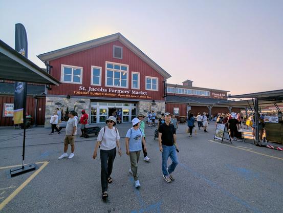 Two storey red building in the background, back lit by the sun, says, "St. Jacob's Farmer's Market." Three people in the foreground, looking to the left and right, and more people milling about behind them.