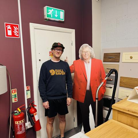 A person stands next to a life-sized cardboard cutout of a man in a red blazer, Mattress Mick. The background features a door, fire safety signs, a vacuum cleaner, and wooden storage units.