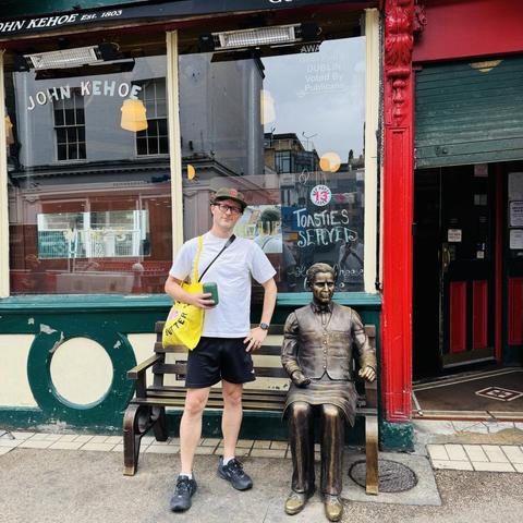 A person stands next to a bronze statue of a seated man on a bench outside a pub named John Kehoe in Dublin. The individual holds a green cup and wears a white t-shirt, black shorts, and sneakers, with a yellow tote bag.