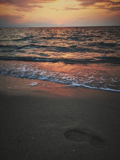 A vertical photo of a sunset over the sea, taken from the shore. The image is divided by the horizon line, with the sky taking up the upper third and the sea and sand the lower two-thirds. The sun, a small orange sliver, is visible on the horizon line. The sky is a warm gradient of orange, pink, and purple clouds. The sea is a calm, dark expanse, reflecting the colors of the sunset. Gentle waves with white foam lap onto the wet sand in the foreground. A single footprint is visible in the sand near the bottom right of the frame. The overall tone is tranquil and warm.