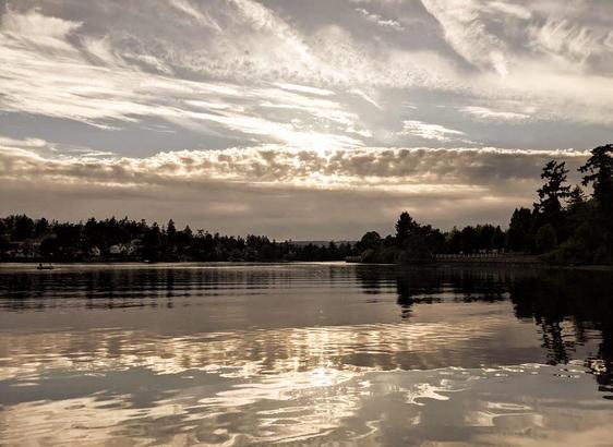 Tranquil waterway landscape with a cloudy sky reflecting in the still water, silhouetted shoreline with trees and homes, and a small boat on the lake.