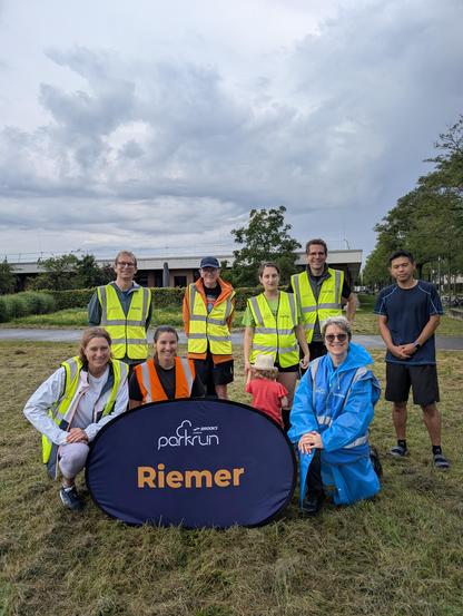 Under stormy skies, volunteers at Riemer parkrun pose for a photo. They wear high-visibility jackets in different colours depending on their roles. The default colour is yellow. The blue jacket is for the Run Director, the orange jacket for the Tail Walker.  They are all smiles. The blue parkrun sign is placed in front of them. They are in the middle of the lawn with a single-storey building behind them.