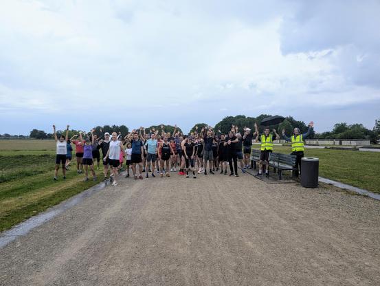 Under stormy skies, runners gather at the starting line of the Riemer parkrun. They raise their arms in the air and seem happy to be there despite the threatening weather. Two volunteers stand on the right, each wearing a high-visibility yellow jacket. One of them will soon give the starting signal.