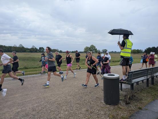 Side view, runners start running. On the runners' side, a volunteer in a high-visibility yellow jacket with an umbrella is standing on a bench. He has his back to us and has just given the start signal.