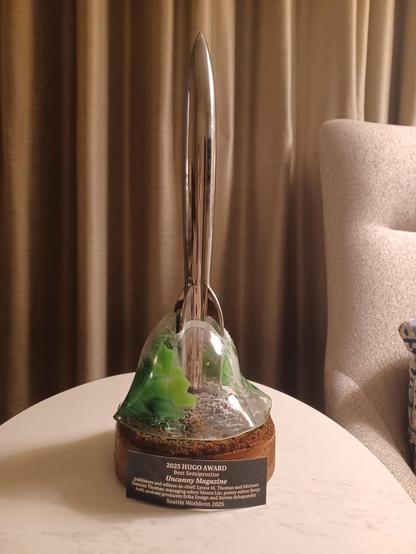The Hugo Award for Best Semiprozine. Chrome rocket on a wooden base with a fragile glass topper swirled with green and snow.