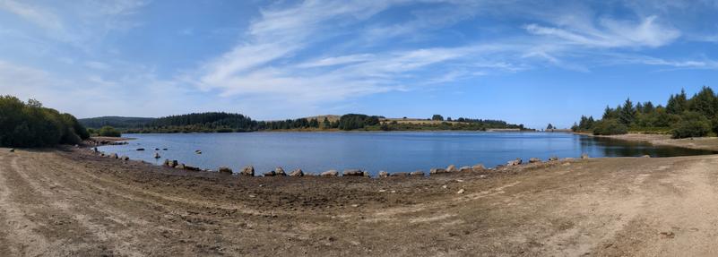 A panoramic view of a calm body of water surrounded by greenery and distant hills under a blue sky with scattered clouds. The shoreline features patches of dry land and rocks.