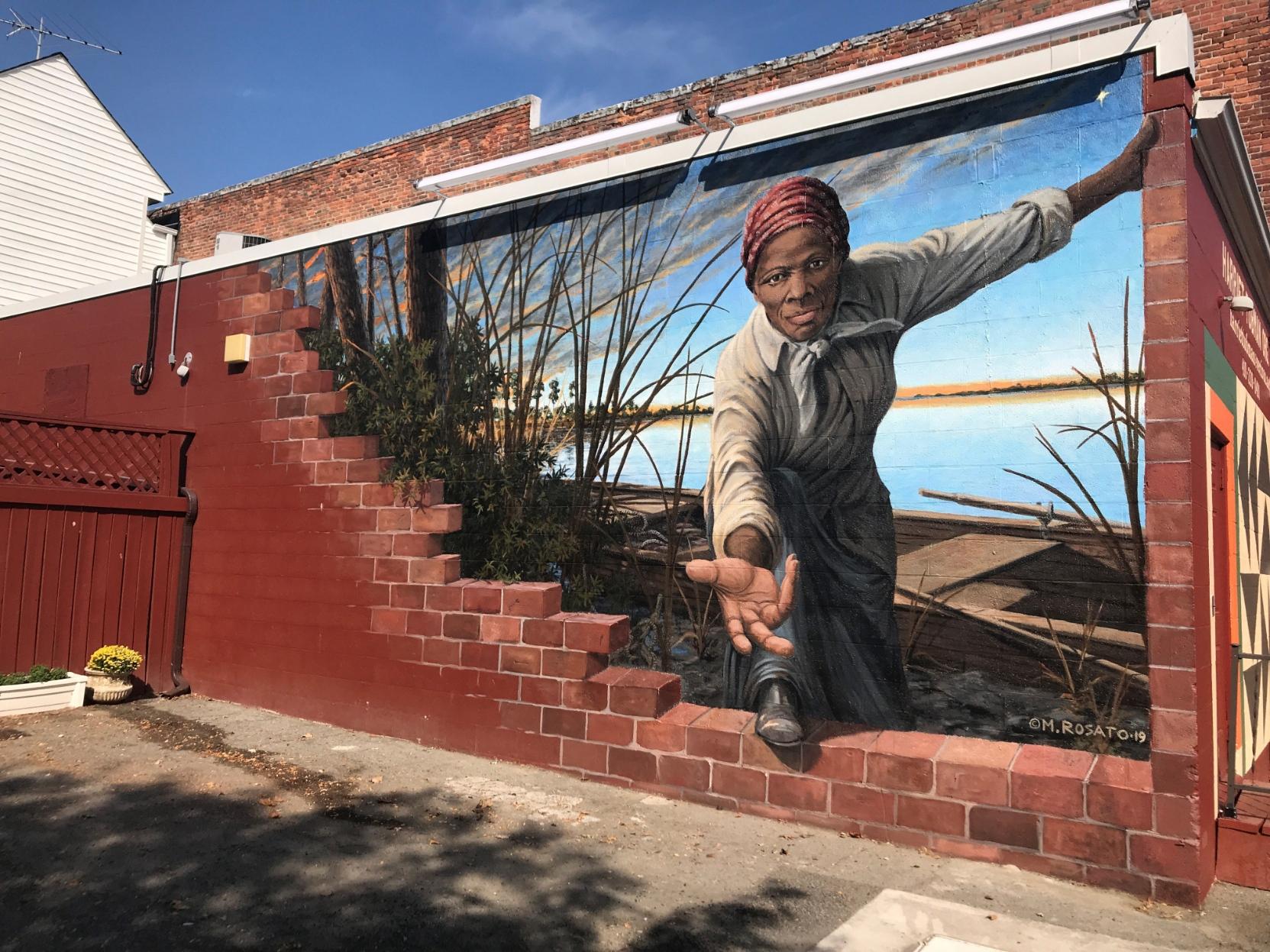 A mural by Michael Rosato on the side of a brick building next to a parking lot. The painting shows Tubman larger than life with blue skies and a river bank behind her, reaching through the wall she's broken through to offer a welcoming hand to those who would step through to freedom.  A mural on brick wall that looks as if the wall is partially crumbled, revealing a black woman reaching through to assist the viewer through the wall and onto a waterway with a rowboat.  https://harriettubmanmural.com/  Harriet Tubman mural artist Michael Rosato