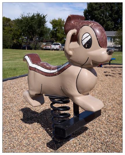 Weathered spring-mounted playground ride in the shape of a cartoonish chipmunk, featuring a brown and cream color scheme with a black base, located on a gravel surface. There are vehicles and trees visible in the background.