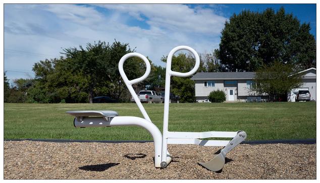 Outdoor playground equipment resembling a large white scissors with a seat attached to one handle and a pedal mechanism on the other, installed on a gravel surface.   It is a manual digging machine, backhoe like.   In the background, a green field bordered by trees and a residential area with houses and parked cars can be seen under a partially cloudy sky.