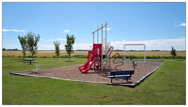 Outdoor playground featuring a red slide, blue benches, and various climbing structures on a sand-filled area, bordered by grass and a few young trees, with a wide open field under a clear blue sky in the background.