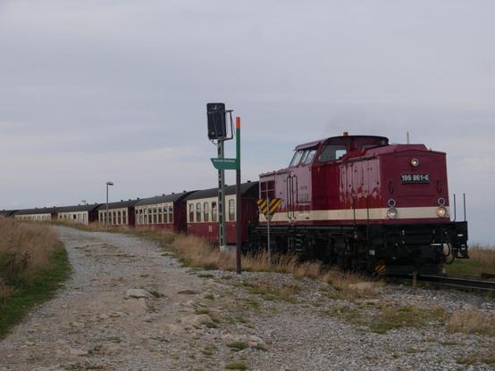 Eine rote Diesellokomotive der Baureihe 199 der Harzer Schmalspurbahnen zieht einen Zug aus historischen Personenwagen aus dem Brockenbahnhof. Sie ist gerade am Ausfahrsignal vorbeigefahren, neben der Bahnstrecke ist der Rundweg auf dem Brockenplateu.